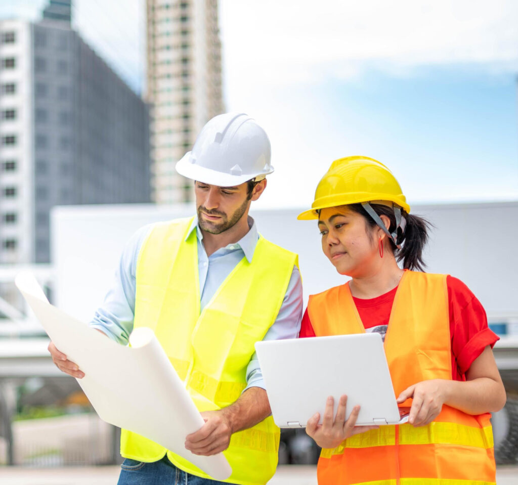 Two construction workers in safety vests review blueprints at a sunny building site.